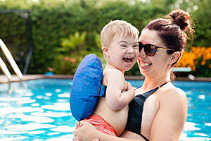 Little boy with Down syndrome having fun in the swimming pool with his mother who is holding him in her arms.