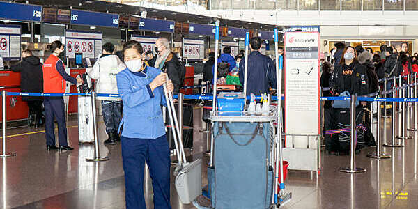 Masked woman in blue cleaning overalls takes a long-handled brush and dustpan from her trolley as passengers queue at the airport check-in area behind her.