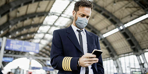 Young pilot in uniform wearing a facial mask looks something up on his mobile phone.