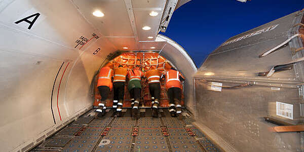Rear view of five men in orange safety vests pushing cargo onto an aircraft.